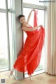 A woman in a red dress is posing by a window.