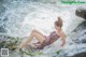 A woman in a pink dress sitting on a rock by a waterfall.